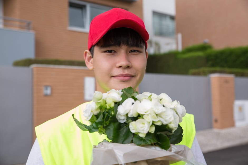 A young male flower delivery person wearing a red cap and neon yellow vest is holding a bouquet of fresh, white flowers with green leaves, wrapped in white paper. He stands outdoors in front of modern