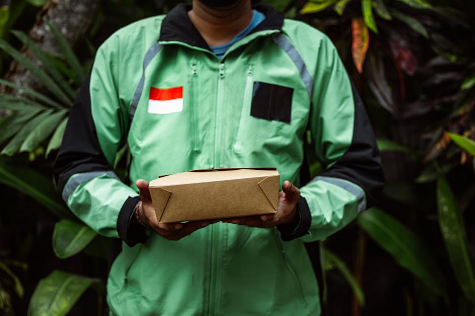A person dressed in a green and black delivery jacket with a red and white flag insignia on the chest, holding a rectangular brown cardboard parcel with both hands, standing outdoors amidst lush green