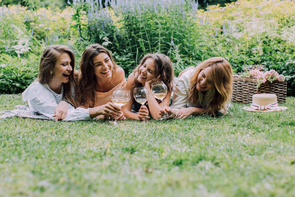 Four women lying on the grass in a park, engaged in a joyful moment with each holding a large wine glass filled with a clear beverage. The women are smiling and laughing, with one supporting her head 