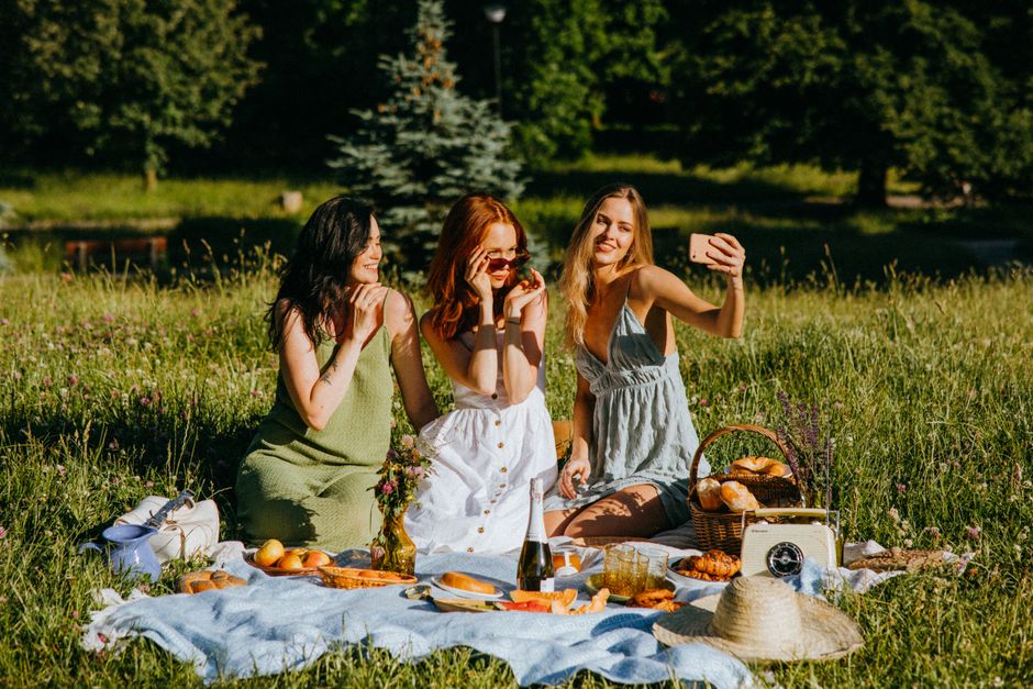 A vibrant outdoor picnic scene with three young women sitting on a light blue blanket in a lush grassy field surrounded by trees, with blooming wildflowers and a small blue spruce in the background. T