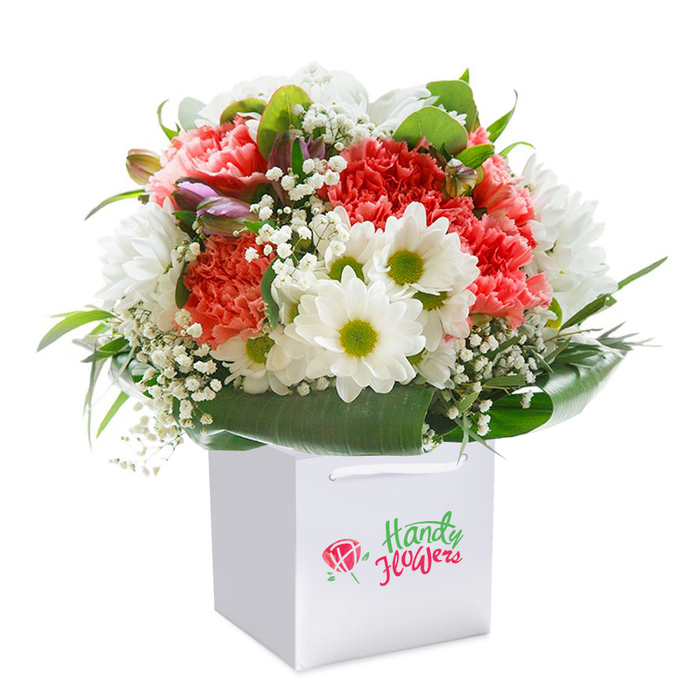 This radiant Brockley bouquet is artfully arranged within a modern, square white gift bag outfitted with practical, elegant ribbon handles-exuding both warmth and sophistication ideal for SE4 celebrations. A lively blend of coral, peach, and deep red carnations, each with soft ruffled petals, creates the heart of the arrangement and infuses it with a rich, textured allure. Interspersed are pure white daisy-like blooms, their vivid lime-green centers adding a pop of brightness and charm evocative of sunny weekends in Brockley's open green spaces. Wispy sprays of delicate baby's breath appear throughout, their airy blossoms lending an ethereal quality and echoing the tranquil beauty of Ladywell Fields. Clusters of closed purple buds add subtle depth and anticipation near the periphery, promising future blooms. All these elements are framed by broad, glossy green leaves that gently curve around the entire bouquet, underscoring the rounded, abundant silhouette. Bathed in gentle, even lighting and set against a flawless white background, every petal and leaf is distinctly visible. This hand-tied bouquet-assembled with the attention and artistry characteristic of Brockley Florist-embodies freshness, elegance, and heartfelt intent, making it a fitting choice for birthdays, anniversaries, or any heartfelt occasion in and around Brockley and Crofton Park.